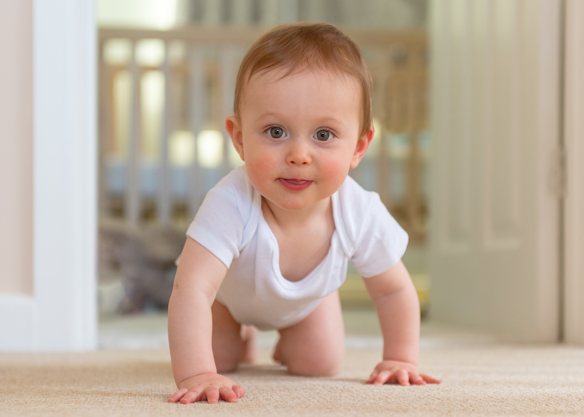 A baby with light brown hair and a white onesie crawls on a carpet toward the camera.